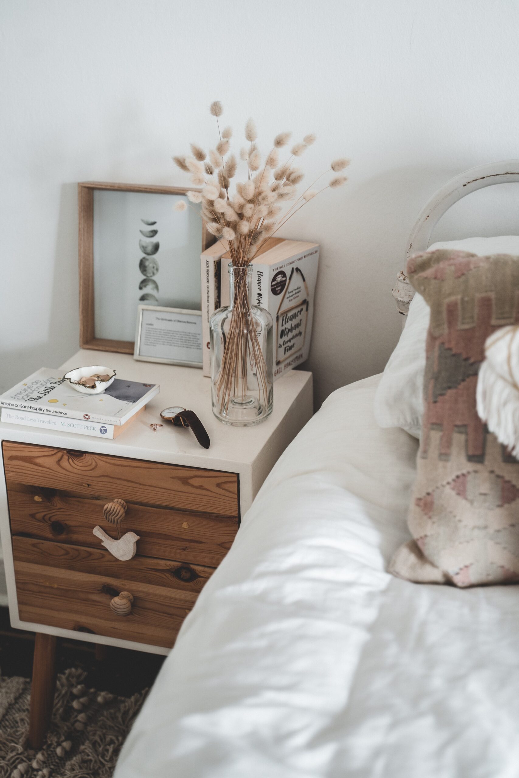 Calm bed with a farmhouse style bed side table containing books, a watch, a vase with dried flowers. 